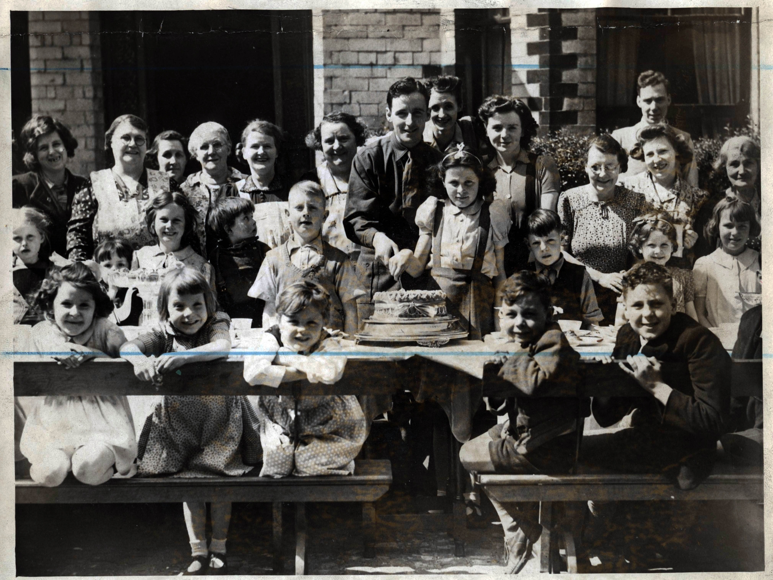 Black and White image of children and young people with military personnel celebrating VE day sitting at a long table with cake during a street party.