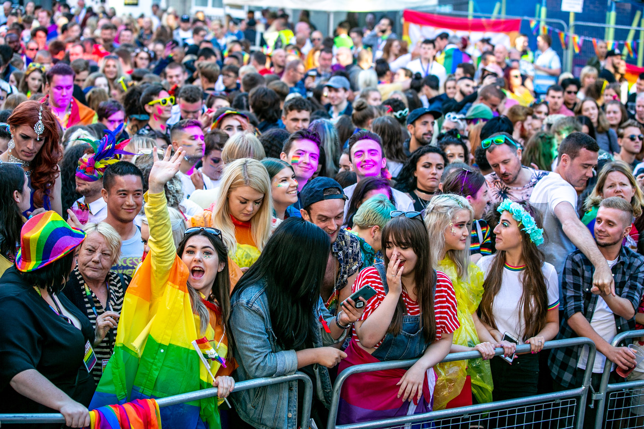 PRIDE festival in liverpool featuring people in a crowd in bright coloured clothing holding rainbow coloured flags.