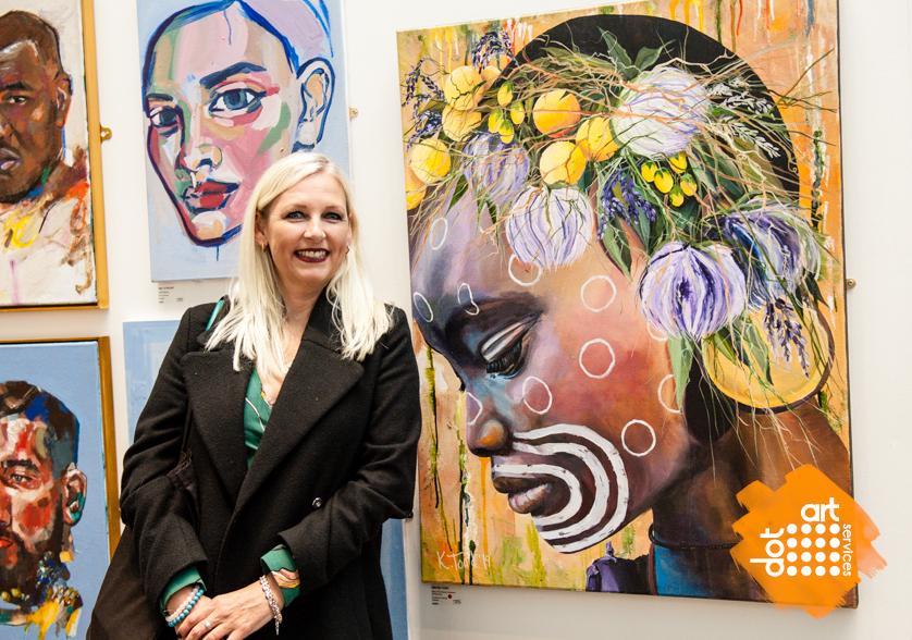blonde lady wearing a black jacket and green shirt standing in front of a canvas taking up the whole background featuring an artistic piece of a head with yellow and green circles on it