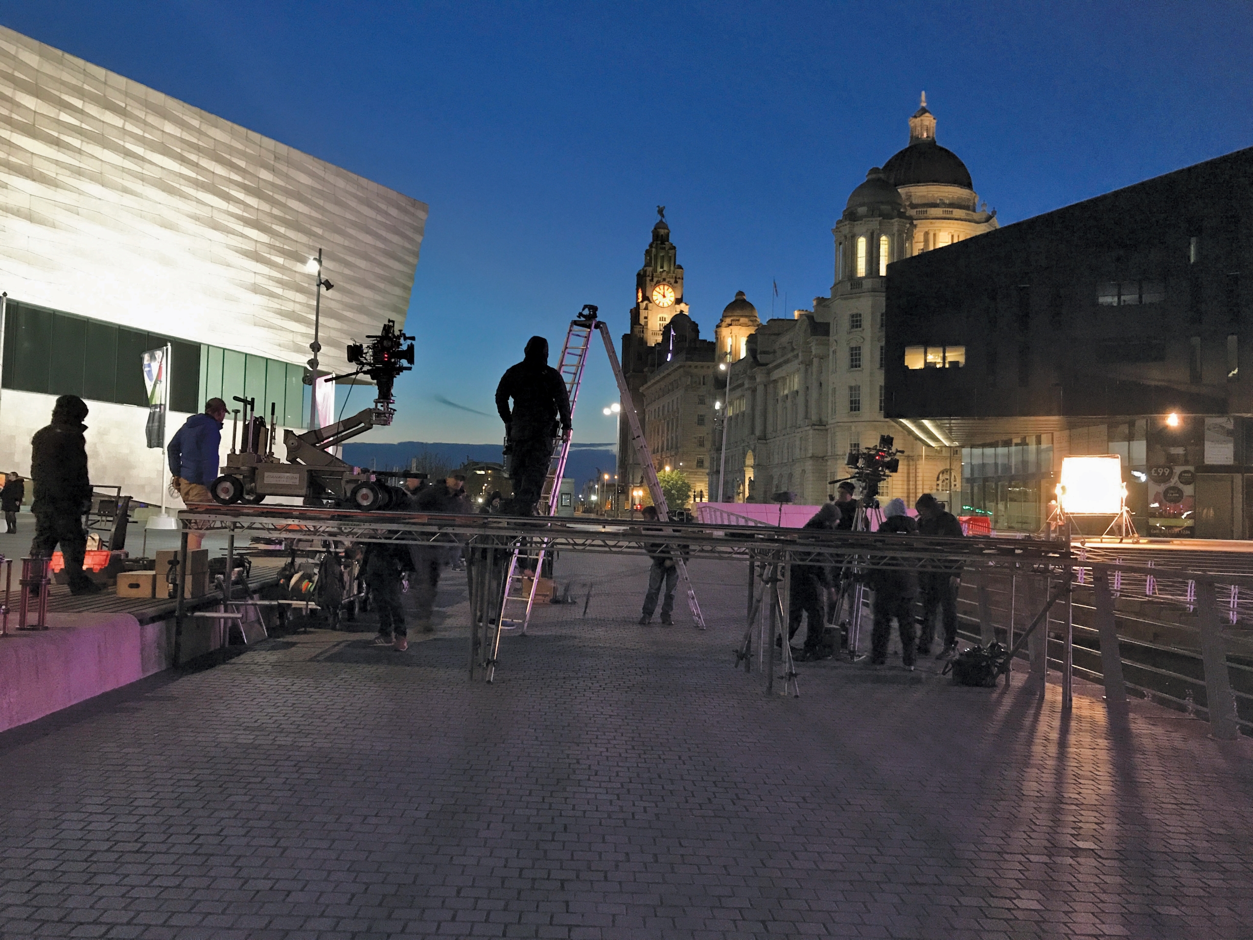 Liverpool waterfont in early evening, film crew filming outside of Liverpool Museum on the flat grey paving and the waterfront buildings are lit up in yellow against the night sky behind the film crew.