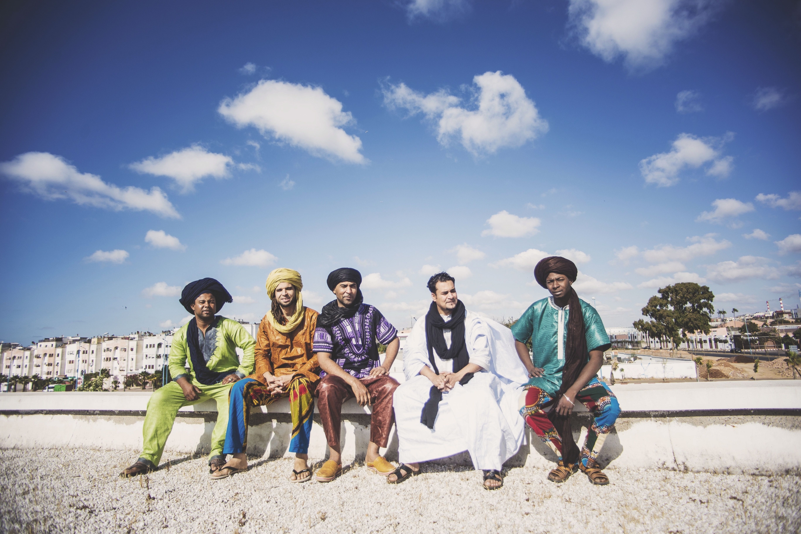 Five gentlem sitting on a white wall in front of a blue sky with white clouds all wearing traditional arabian dress