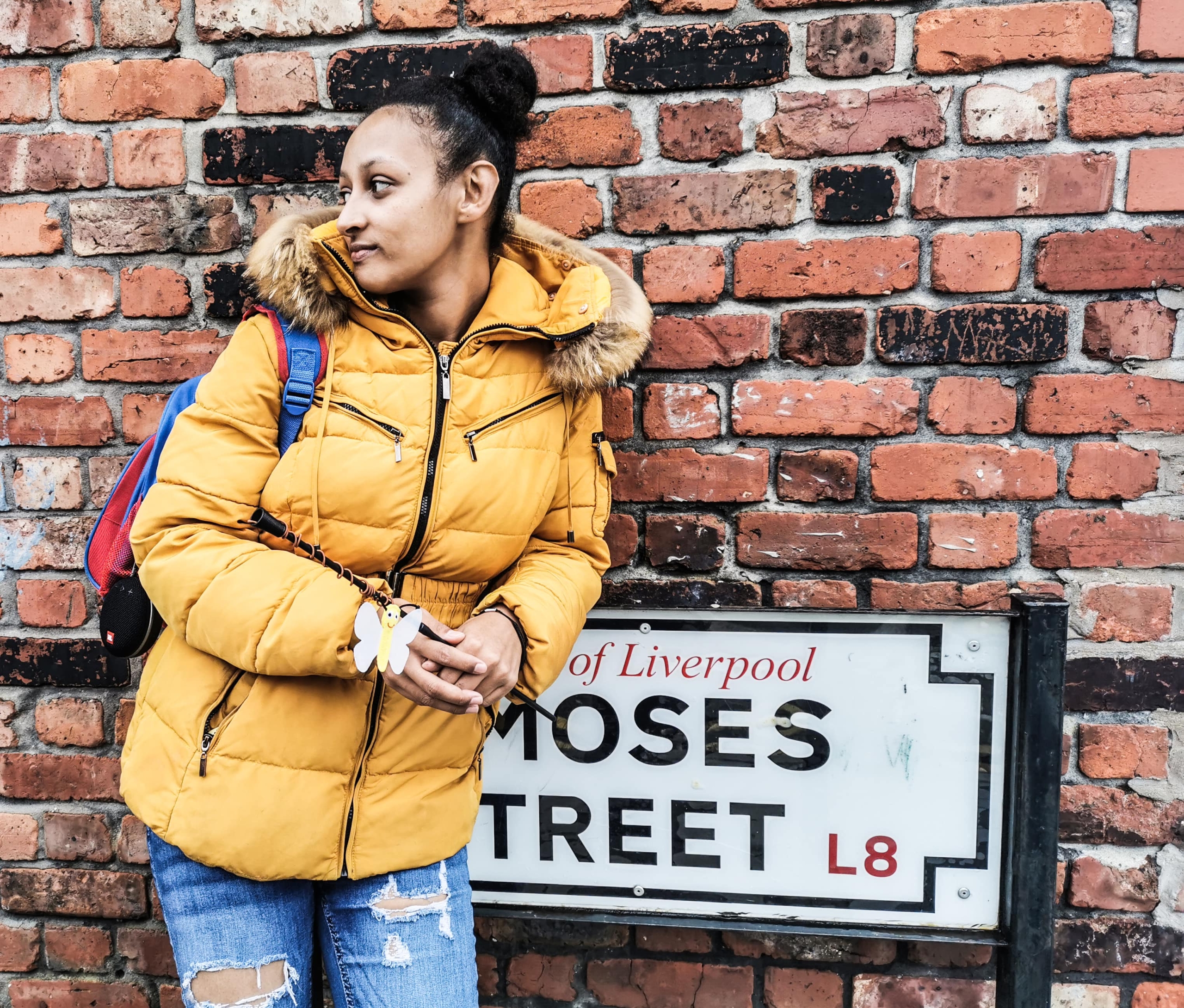 girl in a yellow puffer jacket and light blue ripped heans with a blue backpack stands leaning against a black and white street sign saying L8 and a red brick wall is behind her. Image is copyright to 20 Stories High