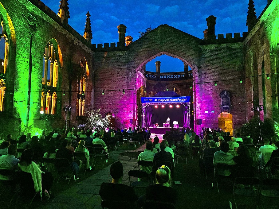 Liverpool Theatre Festival inside the grounds of bombed out church at night with green and purple uplights on the sides of the building and crowds sitting socially distanced on chairs