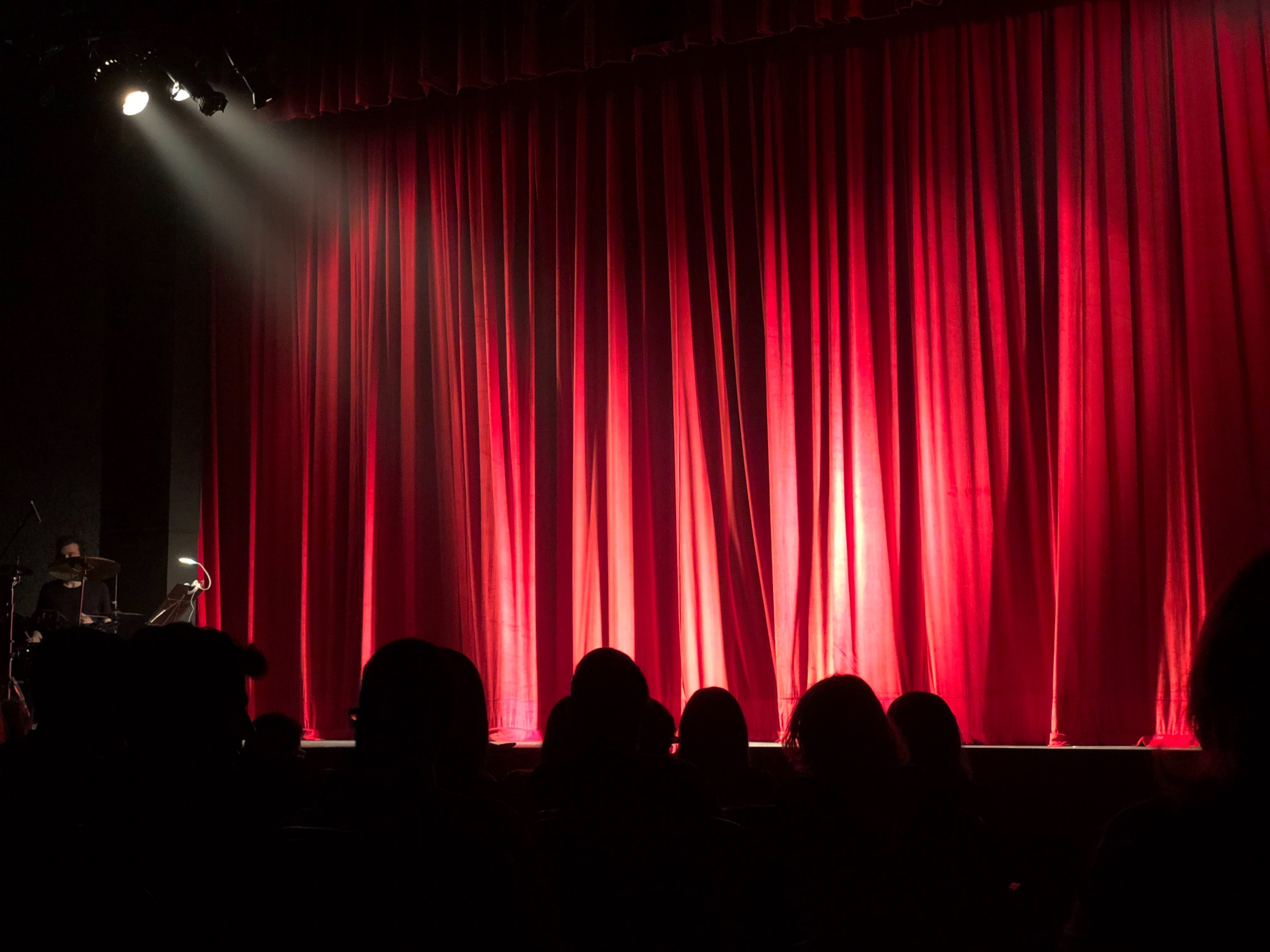 red curtains closed on a stage in a theatre with a spotlight on the stage from the left and the silhouettes of audience members for collective encounters