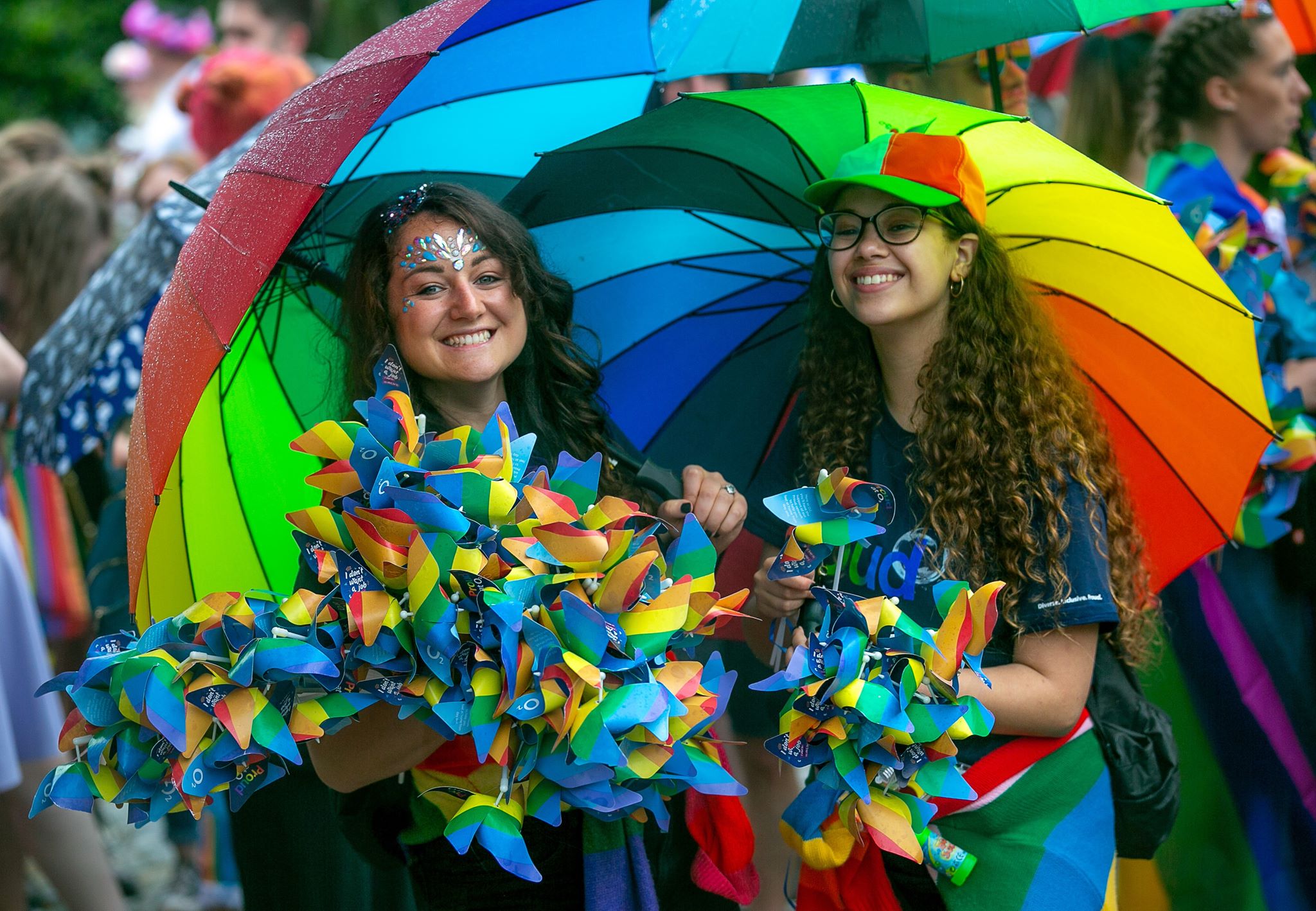 Two girls with long hair, one with face paint, under multi coloured rainbow umbrellas holding multicoloured giant flowers