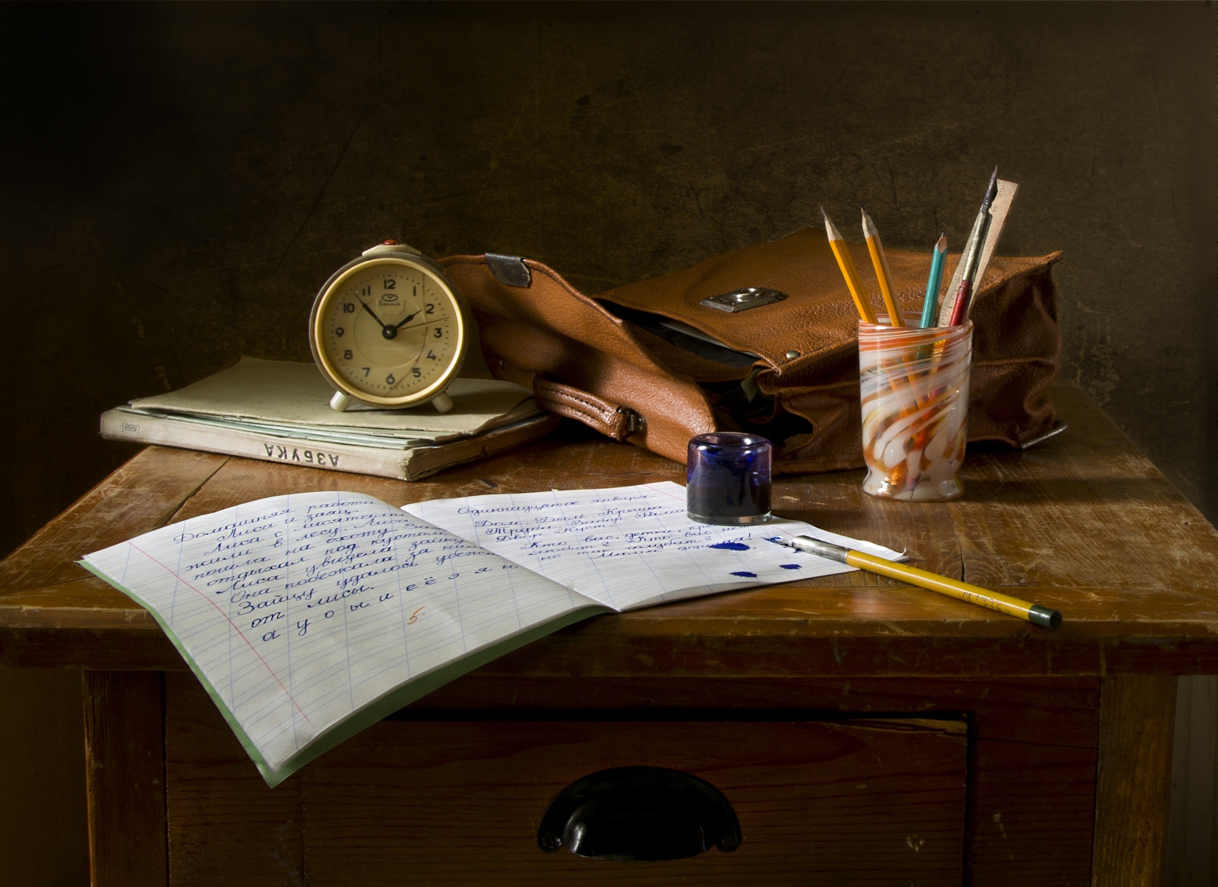 table with a n open notebook, pen pot with pens in a brown bag and a clock used for writing on the wall