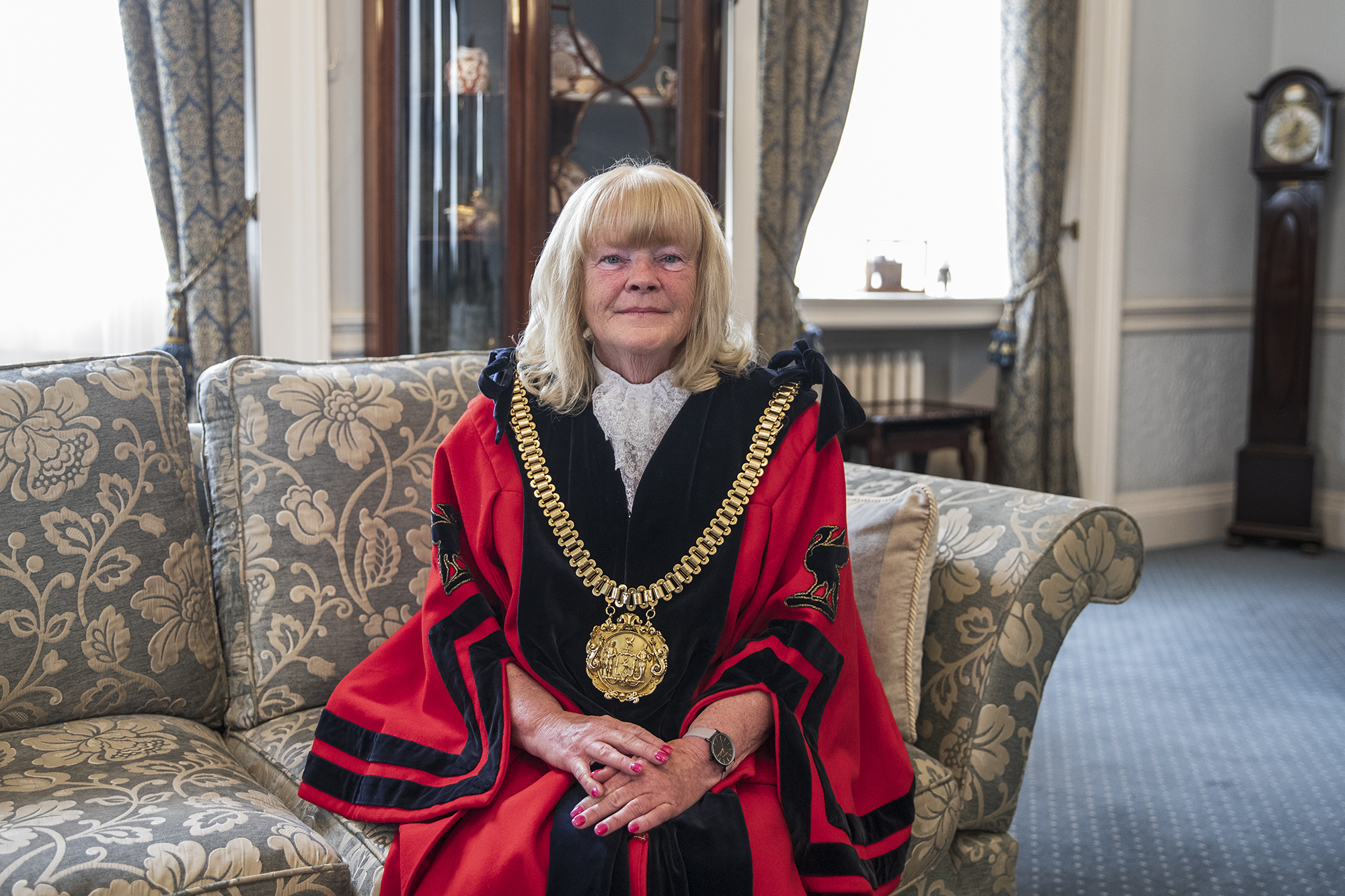 lord mayor mary rasmussen sittin gon a couch in mayoral robes in front of open curtains on a closed window