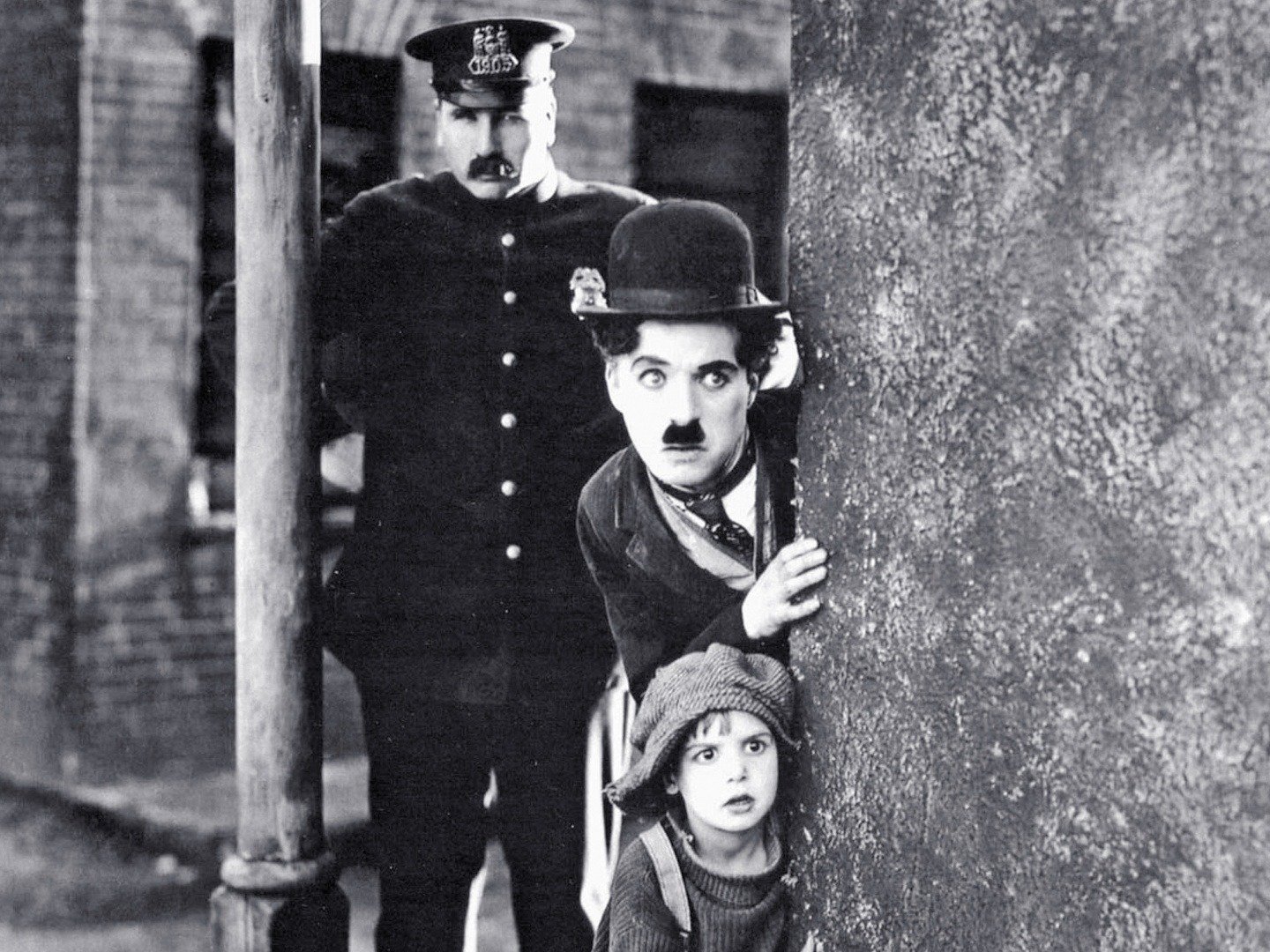 black and white photo of charlie chaplin peering round a wall corner with a small child, a police man stands in the background as part of the angel field festival