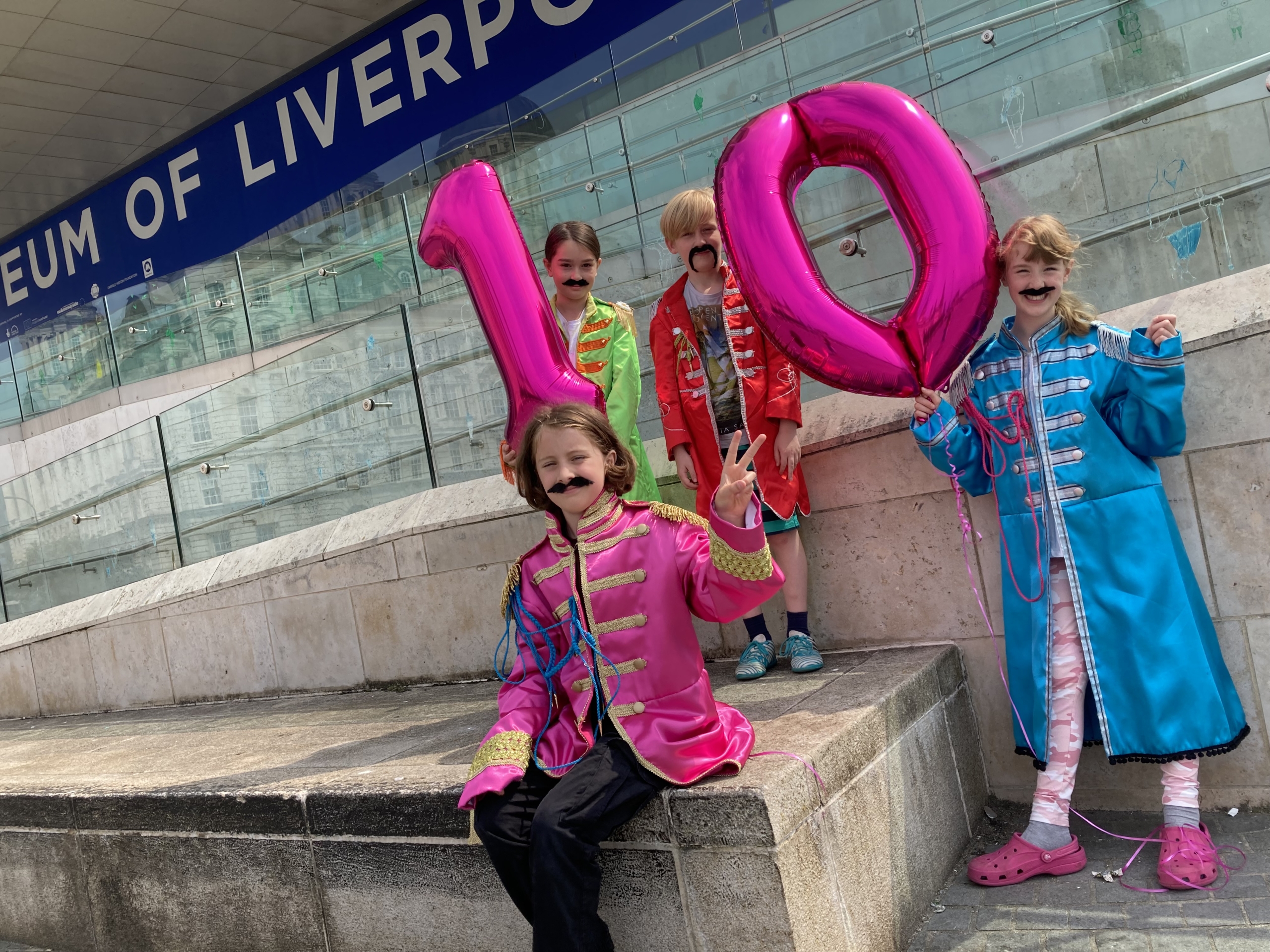 children dressed in beatles costume holding up a pink inflatable ten outside museum of liverpool