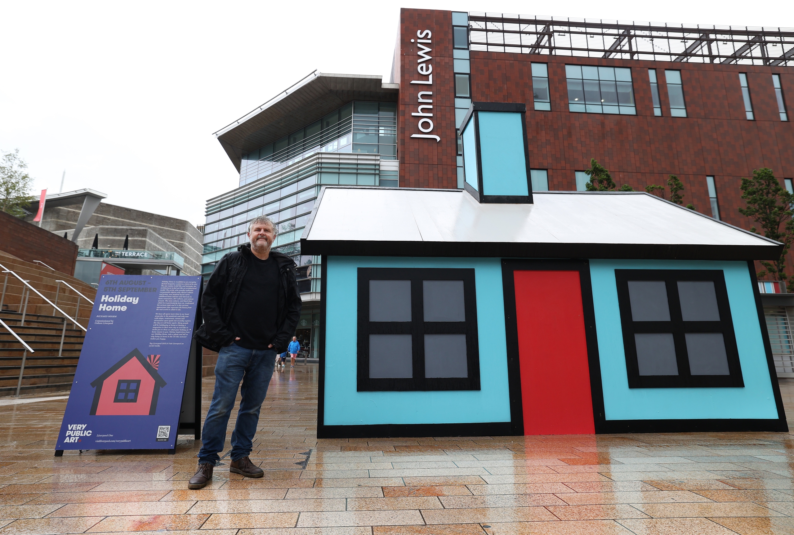 artist stood next to neon blue miniature bungalow called holiday home in Liverpool ONE