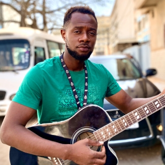 black man playing a guitar in a green t shirt in a street in gambia