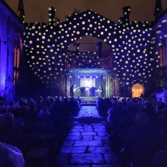 liverpool theatre festival at night in grounds of bombed out church, lighting is blue, view from behind audience looking at a stage with white twinkling lights