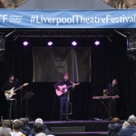 man on stage playing guitar with blue banner overhead says Liverpool Theatre Festival