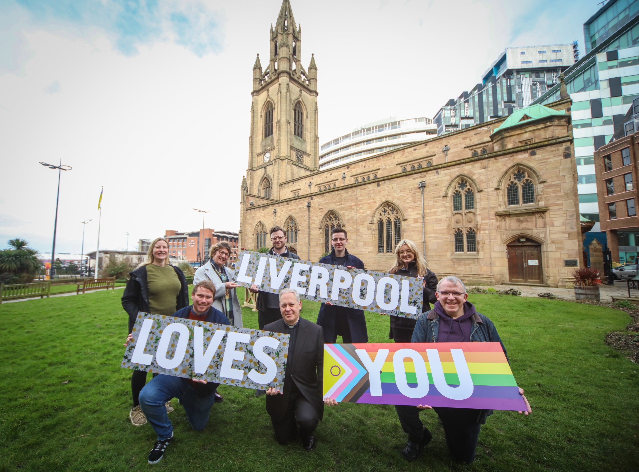men and women standing outside church holding colourful signs that say liverpool loves you
