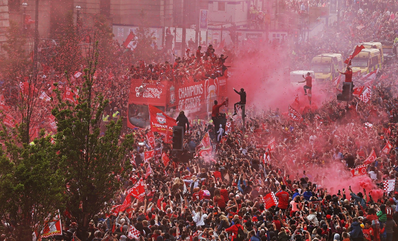 crowds surround an open top bus in 2019 with red flares for LFC Parade