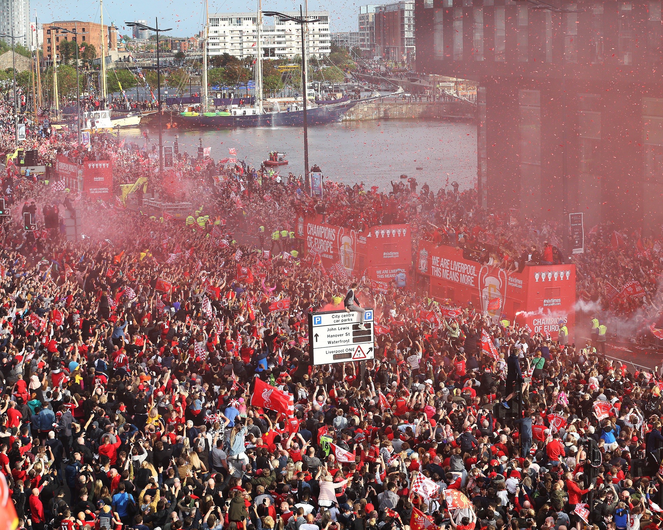 LFC parade from 2019 showing red flare smoke over crowds following two red LFC buses in liverpool city centre