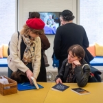 lady showing someone a book sat down at a table with people looking at a screen behind