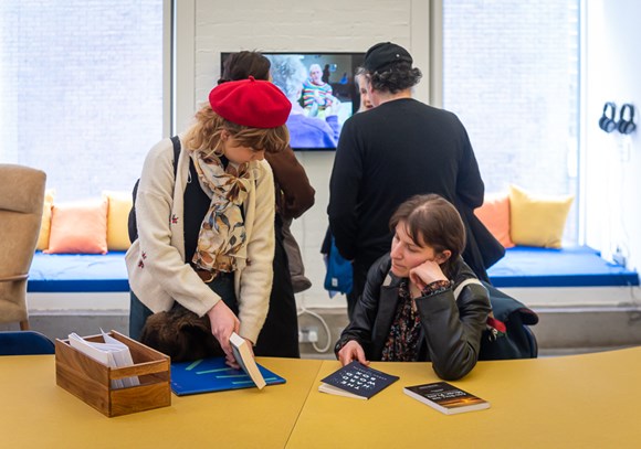 lady showing someone a book sat down at a table with people looking at a screen behind