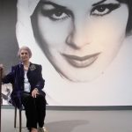 April Ashley sat on chair in front of giant photograph of her in the museum of liverpool