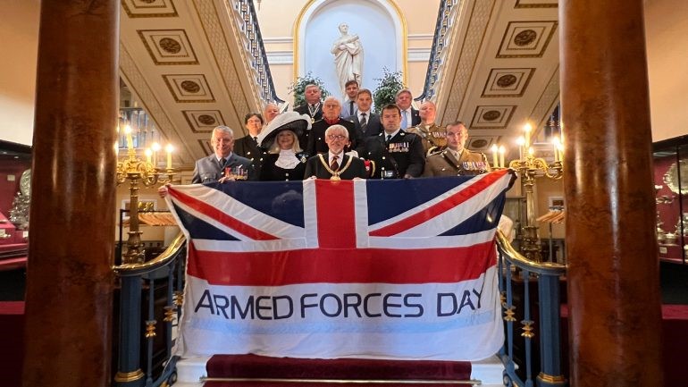 lord mayor and armed forces staff stand on stairs at liverpool town hall celebrating gold award for armed forces holding giant armed forces day flag