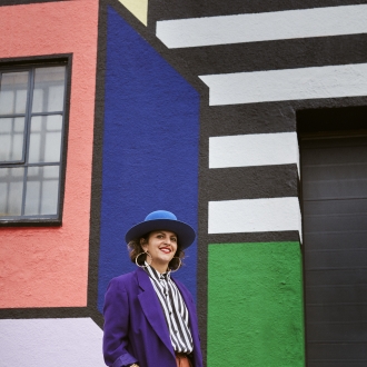 Camille female artist in blue jacket, red pants, blue hat in front of a colourful blue, pink, white and striped wall