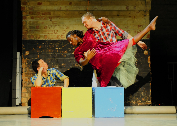 Three actors performing on stage in front of three different coloured boxes