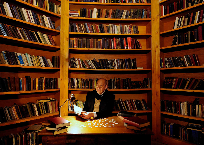 Man sat at a desk with a lamp surrounded by books on shelves