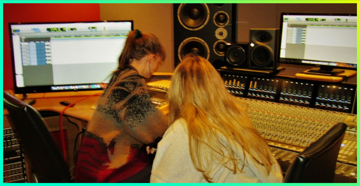 Two young ladies sat in front of a mixing desk, screen and speakers whilst creating music