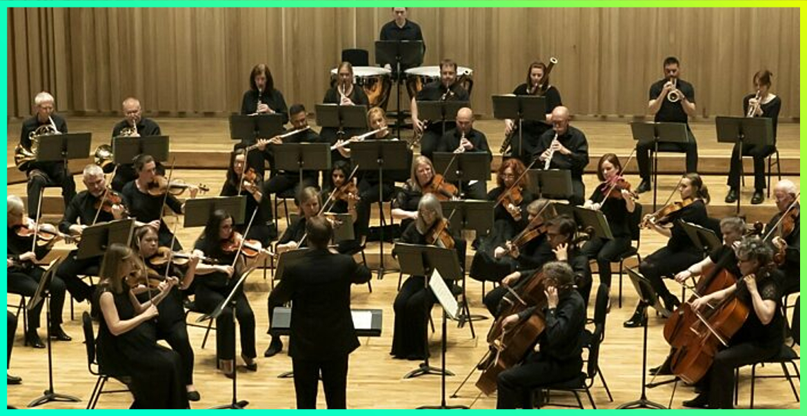 An orchestra of men and women playing various musical instruments are performing on stage, led my a conductor, to a seated audience