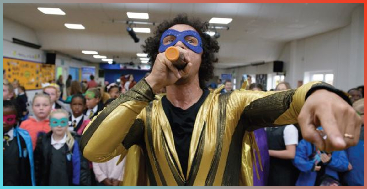 Man dressed up in a gold and blue superhero costume with a blue eye mask to match, holding a microphone with school children looking on in the background