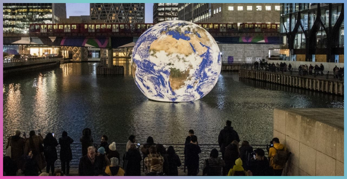 picture of the world floating on water, surrounded by building and people watching on