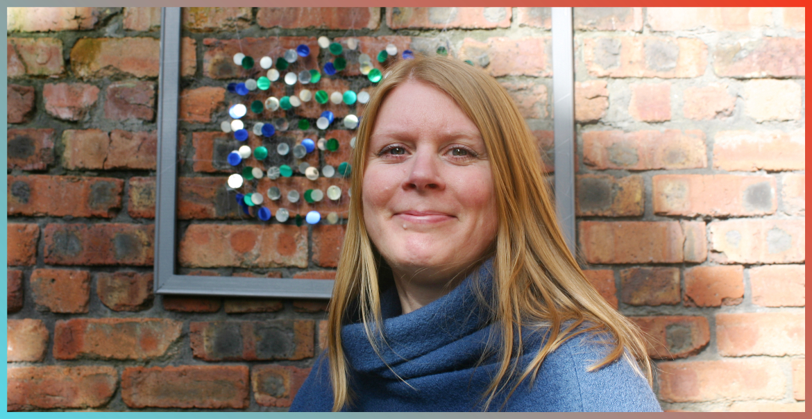 Nicola McGovern smiling wearing a blue turtle neck jumper in front of a brick wall.