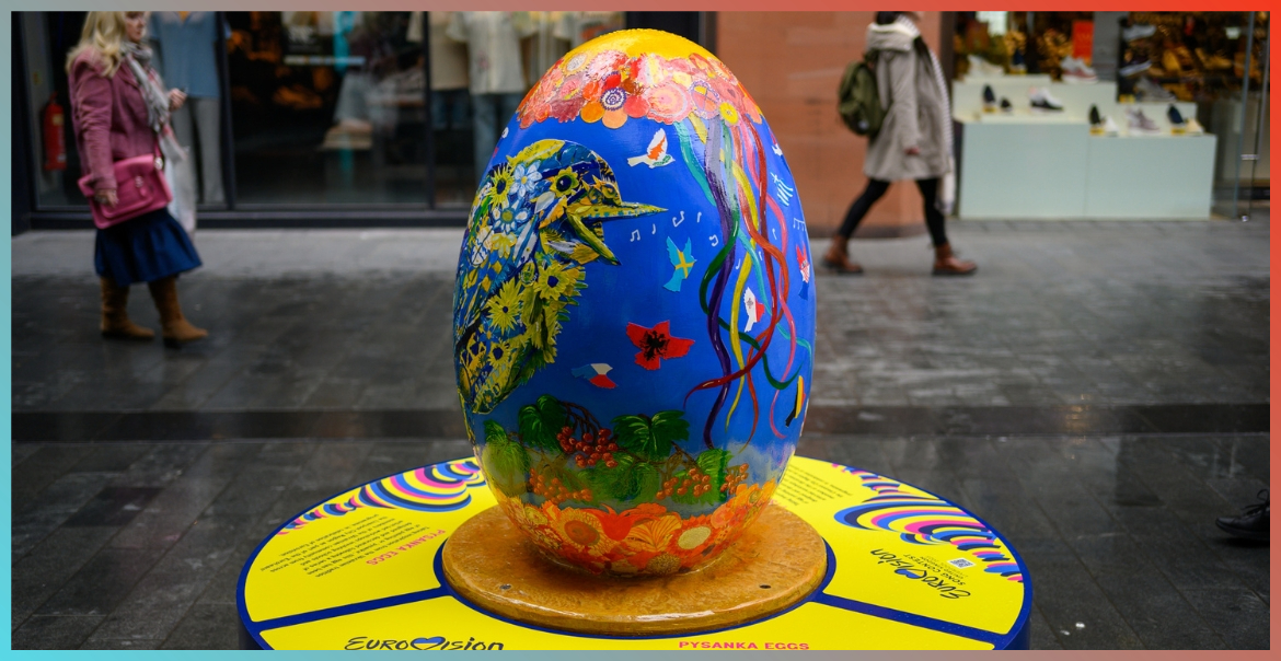 A large egg sculpture painted blue with orange at the top and bottom. In the middle, a bird is created out of blue and yellow coloured feathers. The sculpture is part of a display in the high street,
