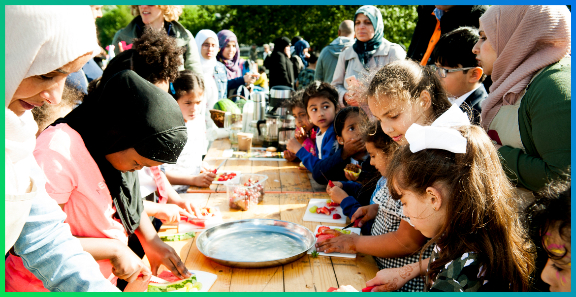a group of children and adults gathered around a table eating different types of food as part of a community event
