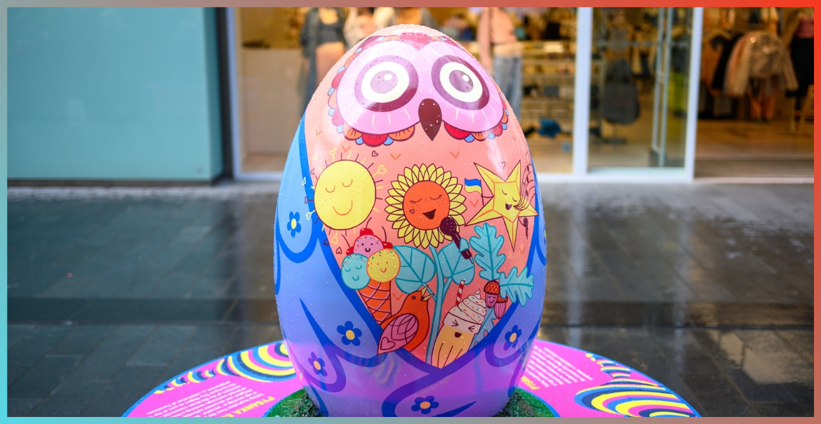 A large egg sculpture with an owl painted on it with pink eyes, blue wings and an orange middle with symbols of the sun, a star, flowers and a milkshake.