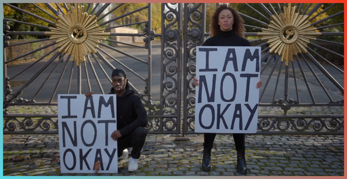 Two people stood in front of ornate gates holding signs saying I Am Not Okay