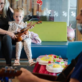 A woman helping a toddler play ukulele while watching a nurse play guitar.
