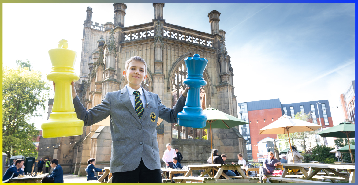 A secondary school student holding up two large chess pieces that are painted blue and yellow. He is in the garden of St Luke's Bombed Out Church in Liverpool.