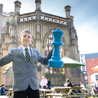 A young boy holding up two large chess pieces, one is blue and one is yellow, in front of St Luke's Bombed Out Church, Liverpool.