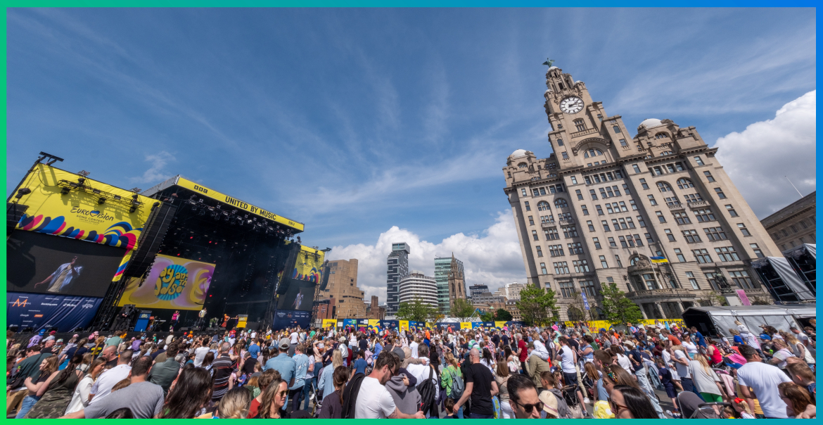 The Liverpool Eurovision Village stage with a busy crowd and the Liver Buildings in the background.