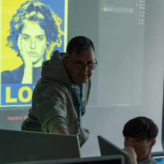 A teacher showing the class how to photoshop an image of a woman on the projector in the classroom while pointing to a pupil's screen.