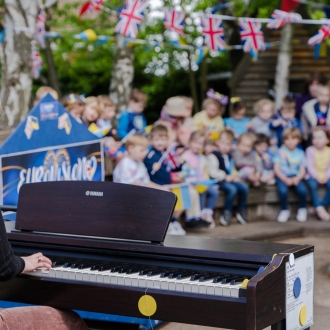 A woman on a piano playing to a class of pre-schoolers outside.
