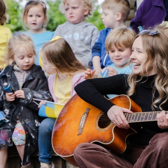 A woman playing guitar to a class of pre-schoolers outside.