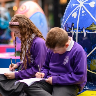 Two children sat next to a large egg sculpture part of the Pysanka Egg display in Liverpool One high street.