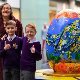 An adult and two children posing for a photo next to a large egg sculpture part of the Pysanka Egg display in Liverpool One high street.