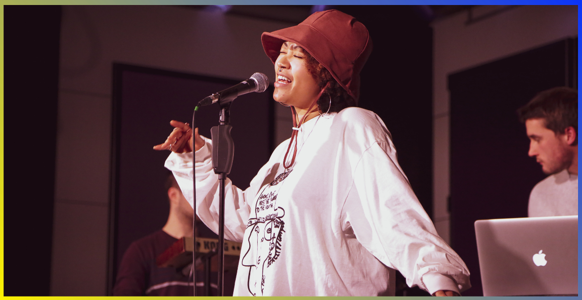 young woman wearing a jumper and hat is singing on stage with a microphone, accompanies by someone on the keyboard