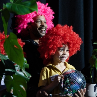 A man and young boy wearing colourful wigs and the young boy is holding a mirror ball.