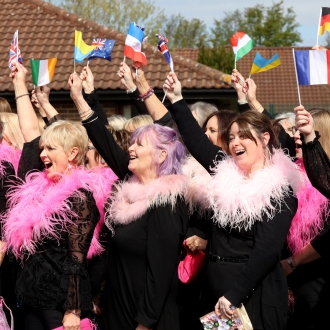 A choir of women wearing pink feather boa's all waving European handheld flags.