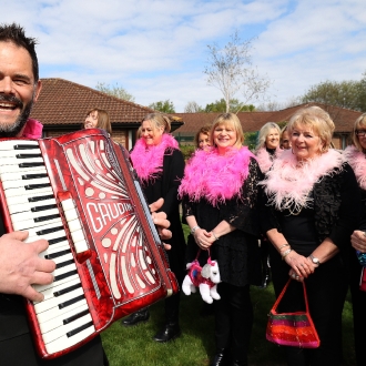 A choir of women wearing pink feather boa's and a man with an accordion.