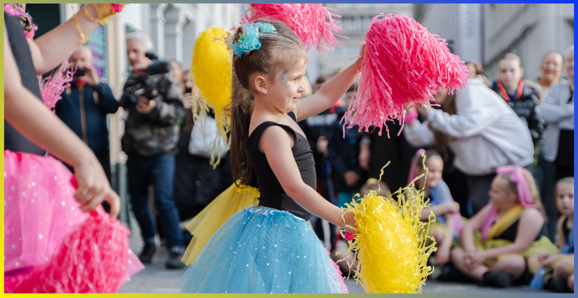 A young girl dancing in a street performance with yellow and pink pom poms.